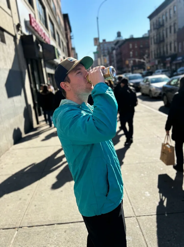 A man in a cyan jacket drinking from a can on a sunny sidewalk