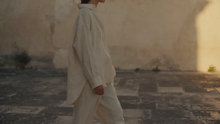 A person in cream linen walks past a stone wall in low golden light