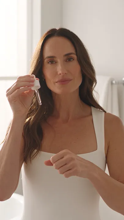 A woman in a bright bathroom holds a serum dropper up to the camera